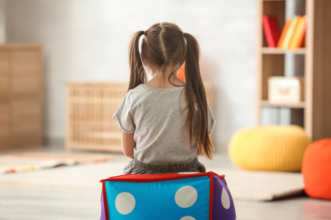 A child, facing away from the camera, sits on a colorful ottoman