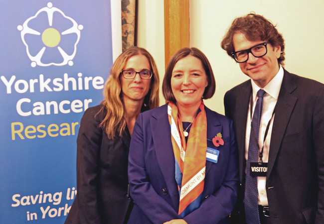 Three people in business attire pose in front of a banner that reads "Yorkshire Cancer Research"