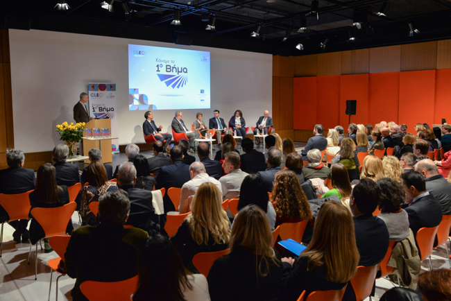 Πολλά άτομα καθισμένα σε ένα χώρο εκδηλώσεων παρακολουθούν μία παρουσίαση.