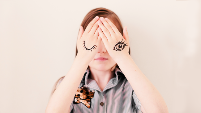 A child holds in front of their face hands with eyes, one open and one closed, drawn on the back of them