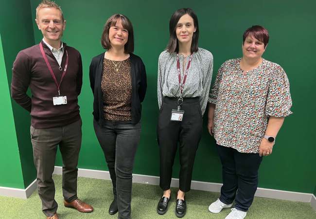 Four individuals smiling pose together in front of a vibrant green wall for a photo.
