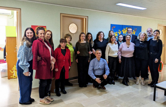 Μια ομάδα από 17 ανθρώπους στέκεται όρθια στον διάδρομο ενός κτηρίου, ο ένας δίπλα στον άλλον, χαμογελώντας στον φακό.