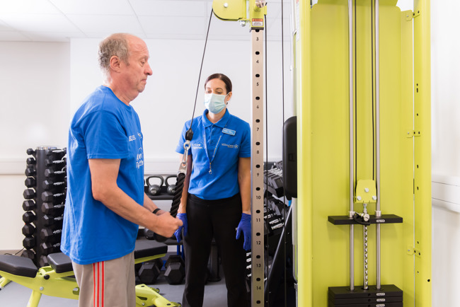 An older adult uses exercise equipment while a trainer in a mask and gloves looks on