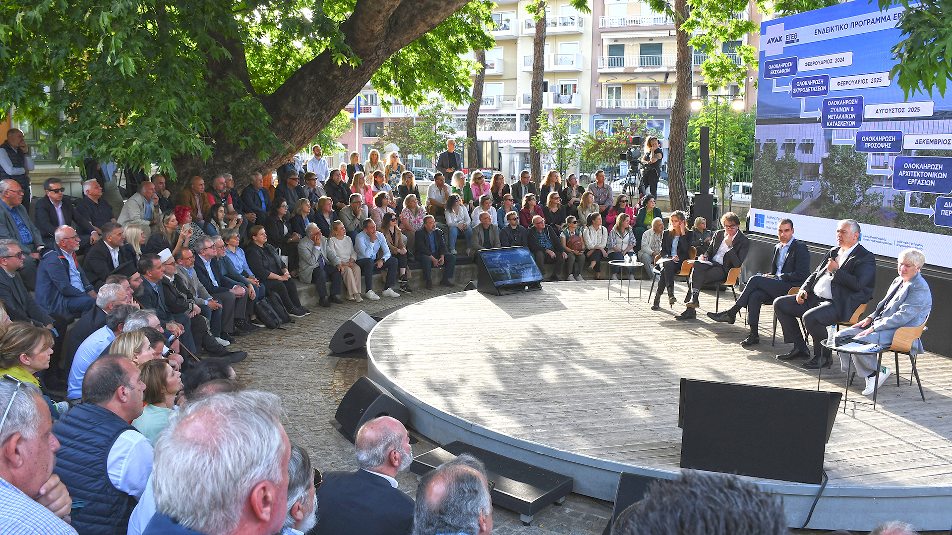 Five people seated on a stage and in front of them there is a diverse seated audience under a tree and they attend the presentation. 