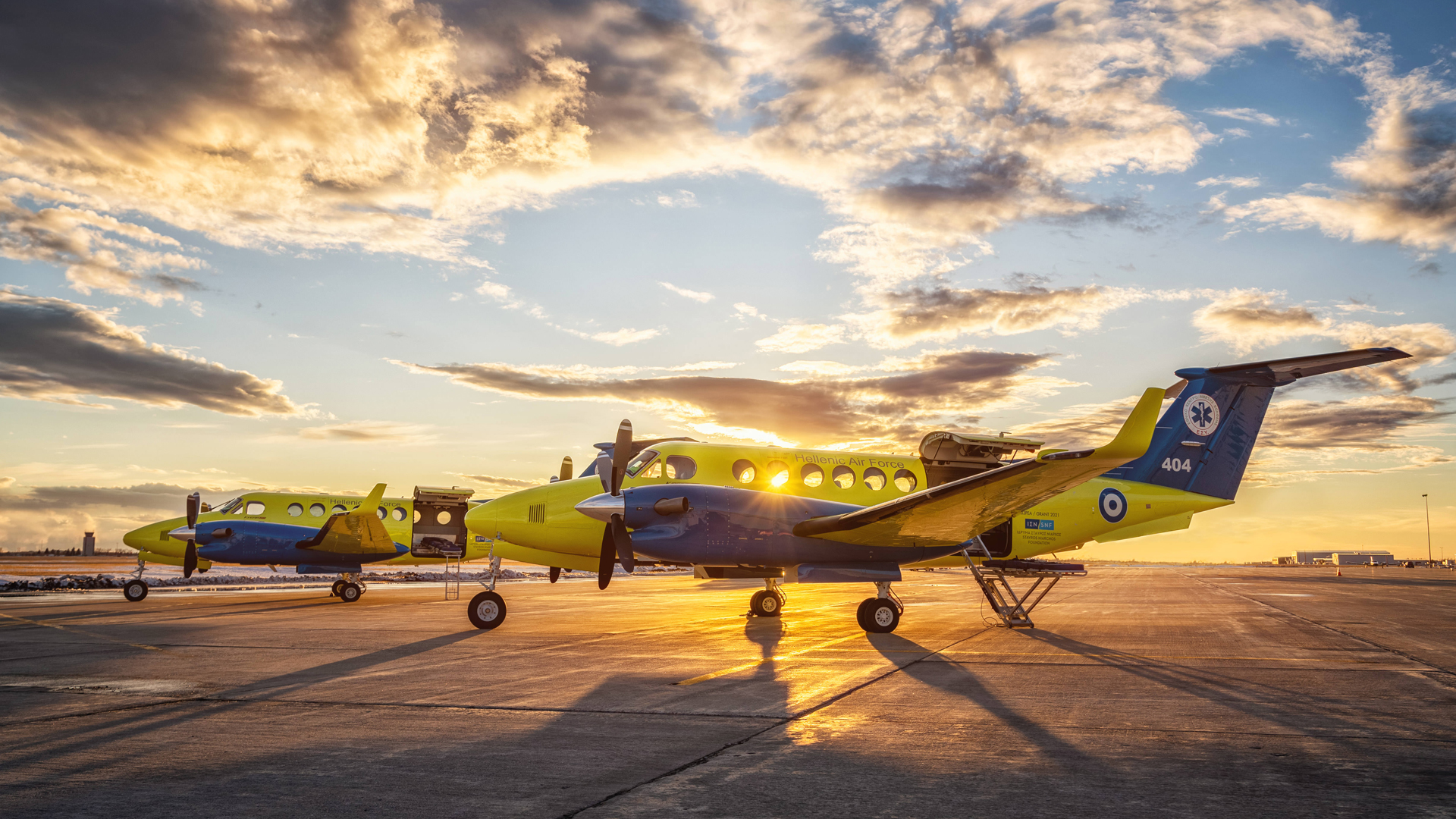 Two yellow and blue medevac propeller planes with the SNF logo and an evil eye sit on the tarmac as the sun sets in the background