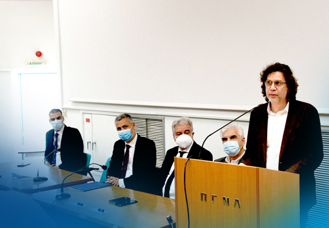 Τέσσερα άτομα με μάσκες κάθονται σε ένα τραπέζι δίπλα σε ένα βήμα στο οποίο μιλάει ένα πέμπτο άτομο