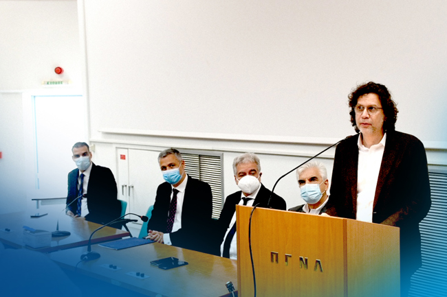 Τέσσερα άτομα με μάσκες κάθονται σε ένα τραπέζι δίπλα σε ένα βήμα στο οποίο μιλάει ένα πέμπτο άτομο